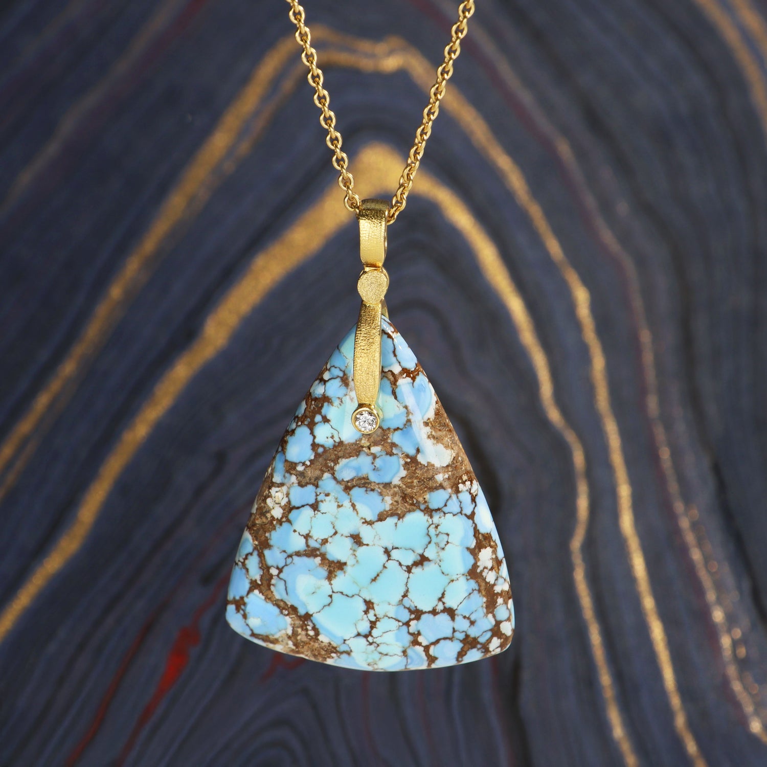 triangular turquoise pendant set with a 18k gold and diamond pendant hanging over a blue and hold background. from Sticks and Stones collection by Alex Sepkus
