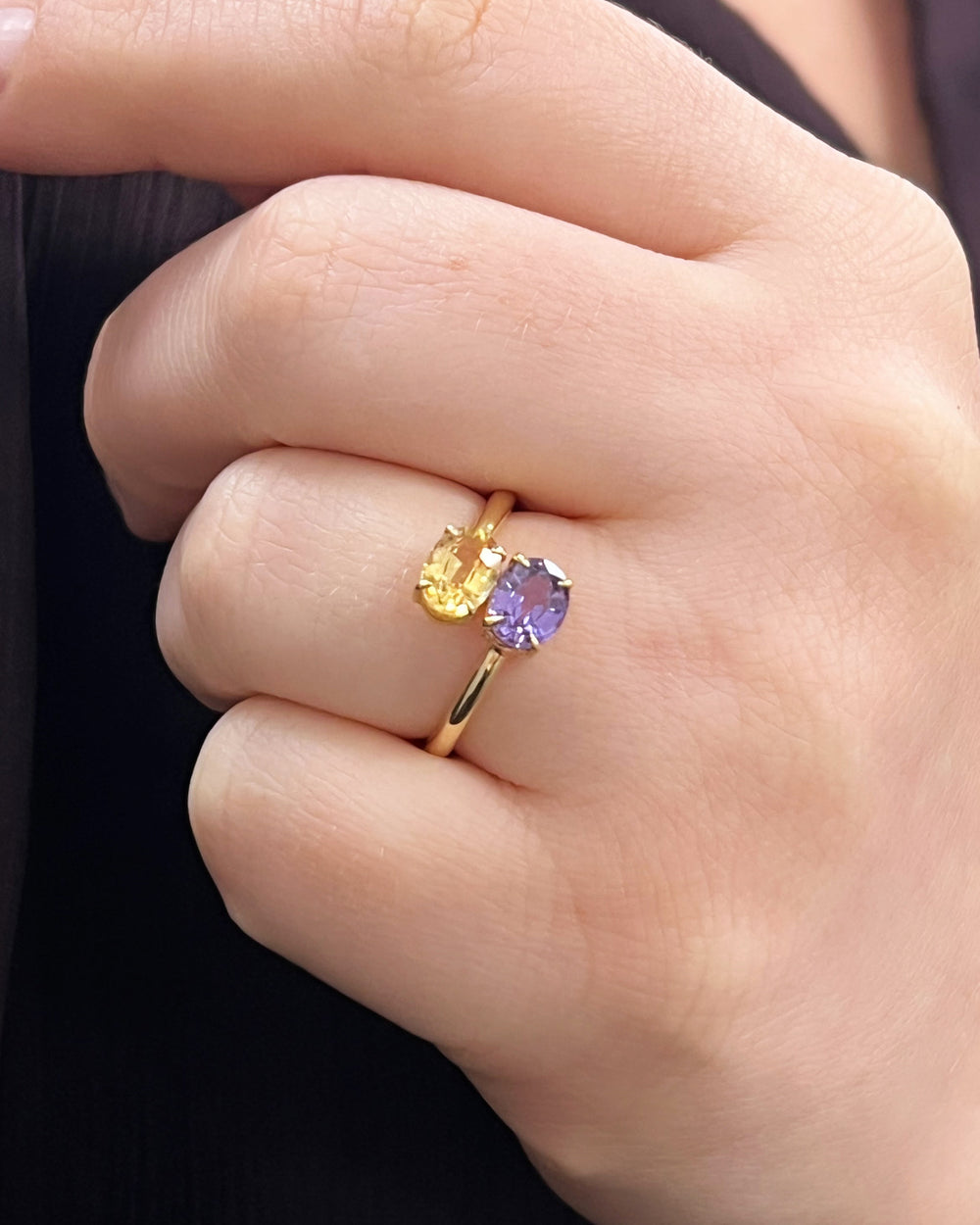 Close-up of a hand wearing a gold ring with two gemstones, one purple sapphire and one yellow sapphire, toi et moi style