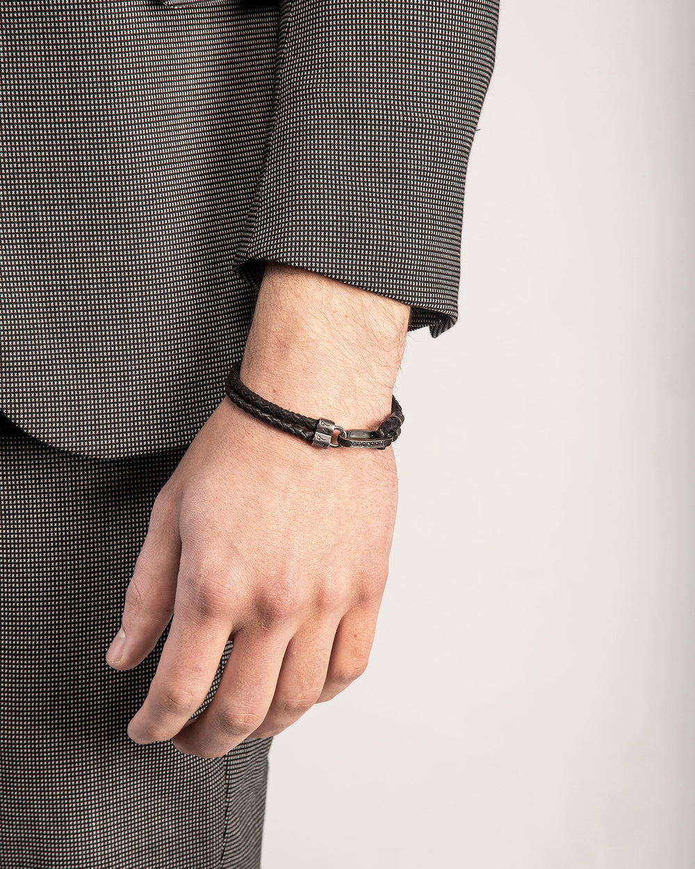 The Parallel Leather Bracelet by Marco Dal Maso is a men’s leather bracelet made from woven black leather with oxidized silver and black diamond accents. Shown here worn with a tailored suit, only the wrist and arm are visible. The bracelet closes with a decorative oxidized silver bar clasp that hooks through a shaped connector, detailed with engraved swirl motifs. It measures 8 1/4 inches in length and is made in Italy. Explore more luxury gifts for men, including leather bracelets, pendants, watches, and