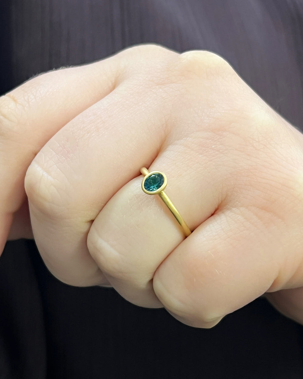 Montana sapphire stacking ring worn on the hand, featuring a 0.44ct oval sapphire bezel set in a dainty 18k gold band, size 6. Style alone or pair with other bands for a personalized stack. Available online or in-store at Green Lake Gallery, located in Seattle and Bellevue. Explore more Montana sapphire jewelry to complete your set.
