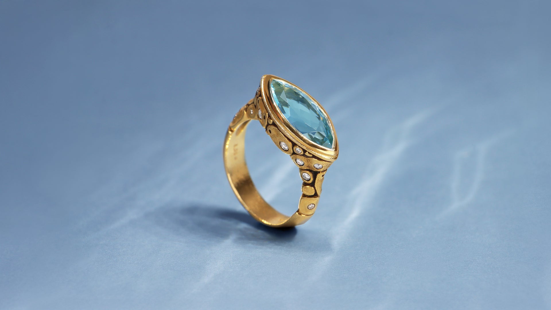 Alex Sepkus design with marquise aqua set in a textured 18k gold ring with diamonds   on a blue background with ripples of light, giving the effect of under water setting