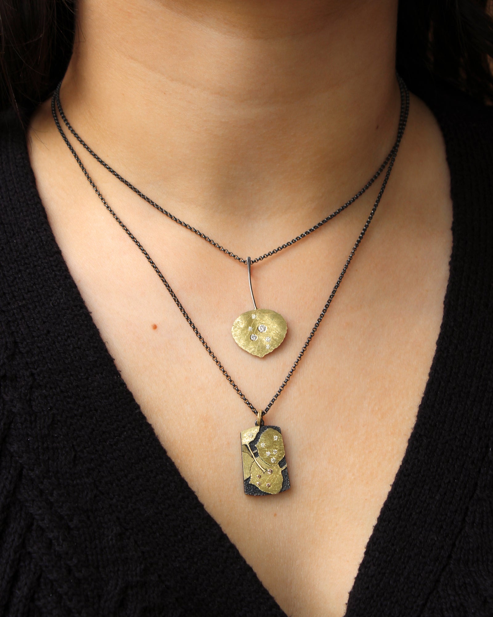 Lady wearing 2 necklaces of contrasting oxidized silver and gold, both featuring nature inspired pendants with gold leaves and diamonds