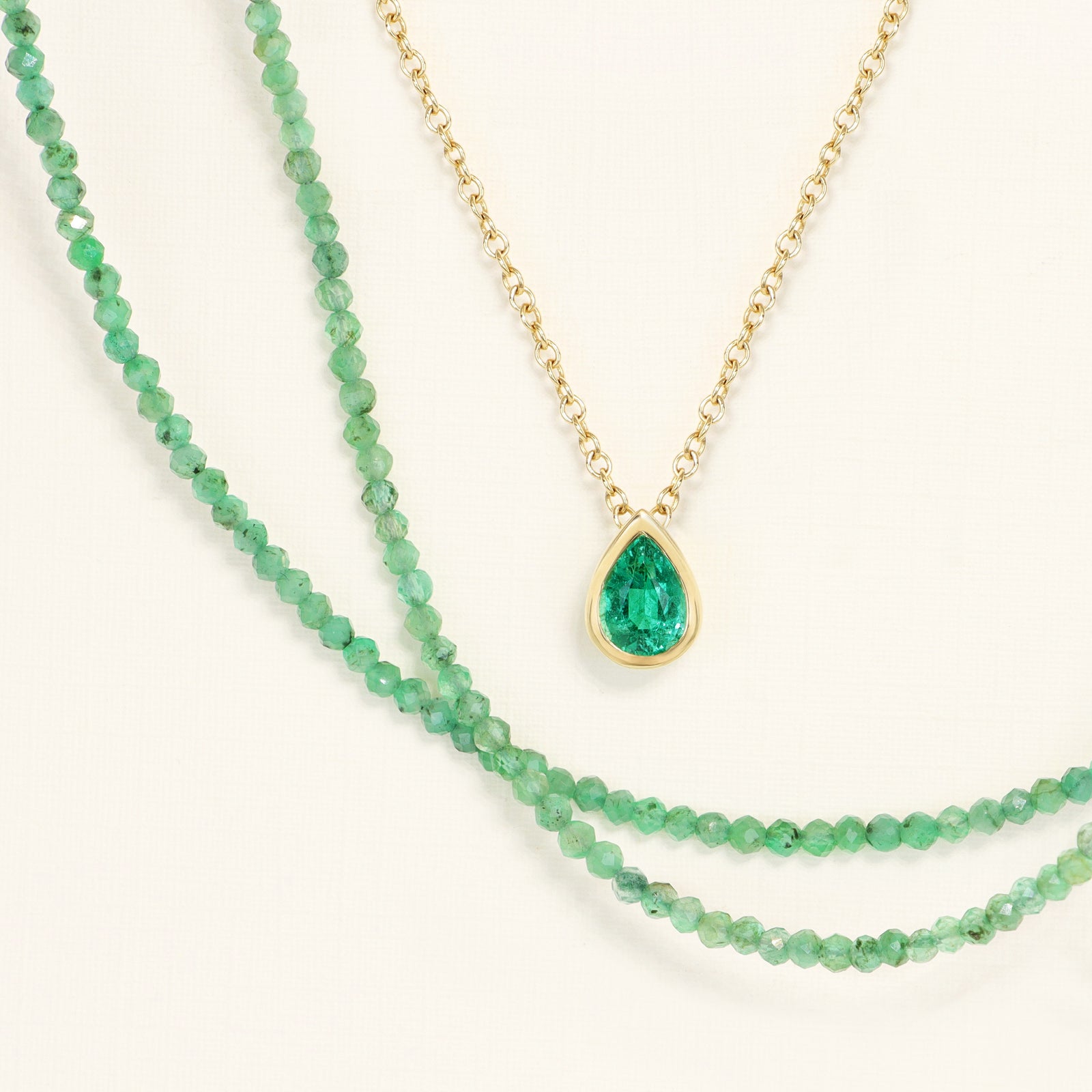 A pendant with a Pear shape Emerald bezel set in 18k gold on a chain and faceted emerald bead necklace on a cream background 