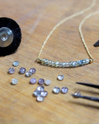 A gold chain necklace with a curved bar of small iridescent beads, displayed on a wooden surface with tiny loose rhinestones, tweezers, and a small black wheel tool.